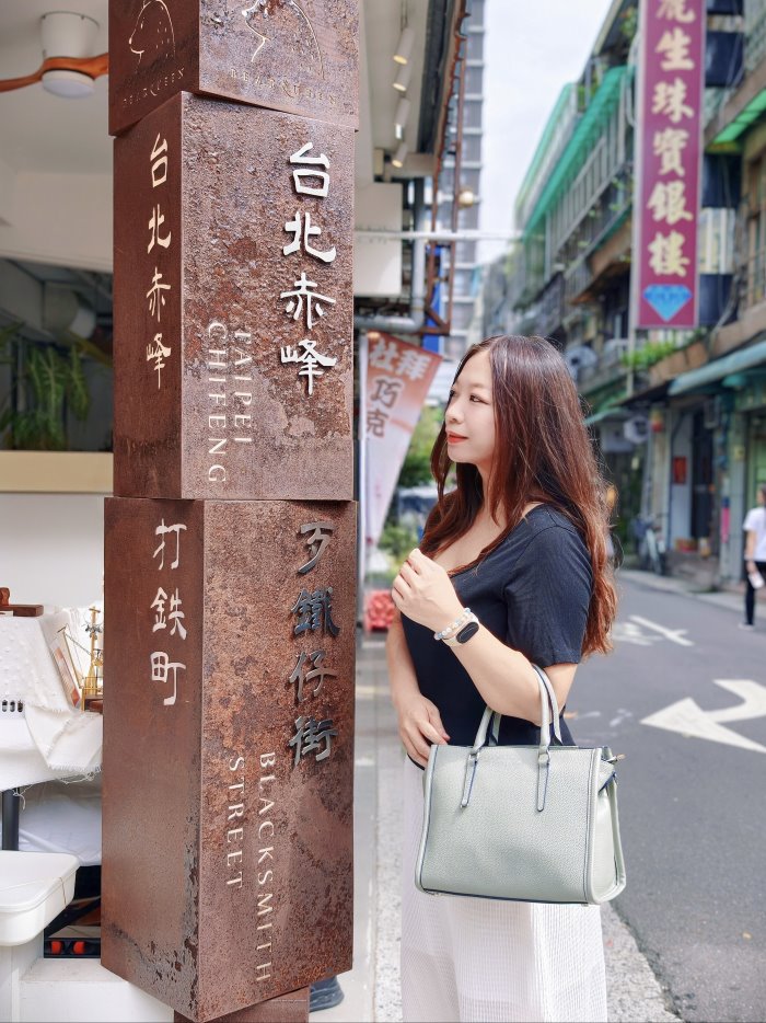 台北赤峰街打鐵仔街生鏽地標鐵柱旁，身穿黑上衣白長裙手挽霧綠方包